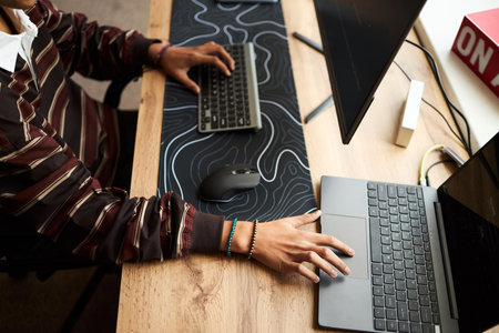 Young adult Black man working at desk using wireless keyboard and laptop, hands typing and navigating mouse, modern workspace setup with dual monitors and office accessories visibleの写真素材