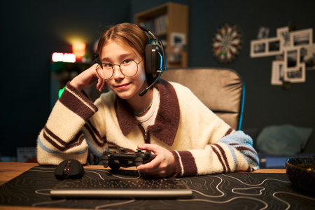 Portrait of Caucasian teenage girl wearing headset sitting at desk holding game controller and smiling into camera, gaming equipment and computer keyboard visible in foregroundの写真素材
