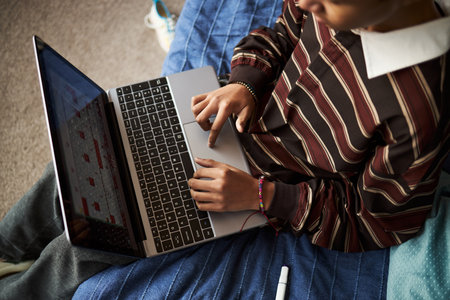 Teenager boy with medium skin tone sitting on bed using laptop, focusing on screen and typing with both hands, wearing striped long sleeve shirt, studying or working remotelyの写真素材