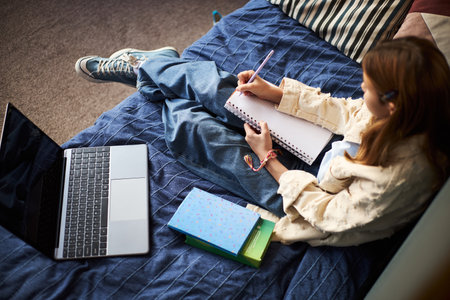 Teenage girl sitting on bed writing in notebook with pen, laptop open beside her, textbooks stacked nearby, studying or doing homework in casual home settingの写真素材
