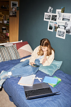 Teenage Caucasian girl sitting on bed using smartphone, surrounded by open laptop, notebooks, and...
