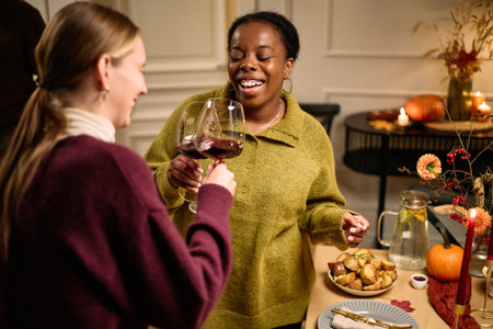 Young adult Caucasian woman and young adult Black woman clinking wine glasses and smiling, while...