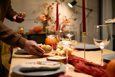 Hands of anonymous man arranging food on festive table with empty plates, wine glasses, decorative pumpkins and candles during Thanksgiving dinner among friends, partial body visibleの写真素材