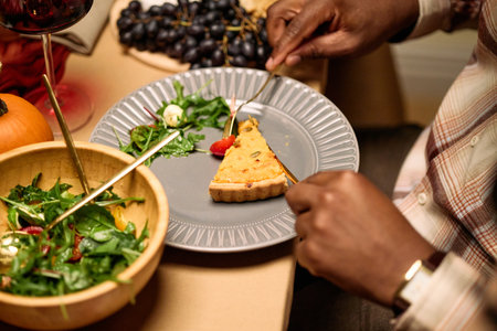 Black man sitting at table eating slice of pie with fork during thanksgiving dinner among friends, holding plate with salad and grapes visible, wearing plaid shirt, hands in focusの写真素材