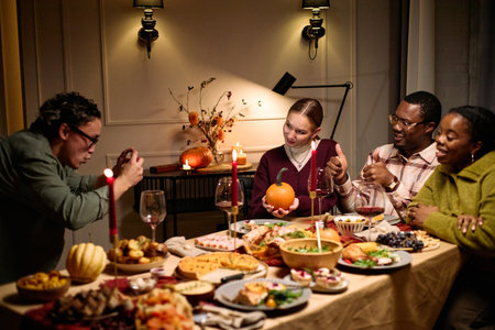 Group of multiethnic friends sitting around table sharing Thanksgiving dinner, smiling and posing while Caucasian man taking photo, Caucasian woman holding pumpkin, food and candles visibleの写真素材