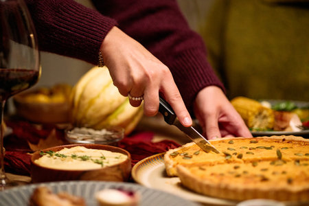 Unrecognizable woman slicing pumpkin pie with knife during Thanksgiving dinner among friends, visible hands and partial arms, sharing food at festive table with assorted dishesの写真素材