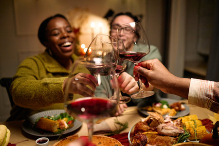 Diverse group of young people and middle aged friends clinking wine glasses while celebrating Thanksgiving dinner, plates with traditional holiday food on table, smiling and enjoying meal togetherの写真素材