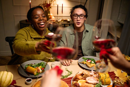 Young adult Black woman and young adult Asian man smiling and raising wine glasses with friends during Thanksgiving dinner, diverse group celebrating around festive table with foodの写真素材