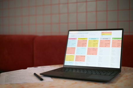 Laptop displaying colorful project management dashboard with development stages for new AI model, resting on table in business setting, printed documents lying beside deviceの写真素材