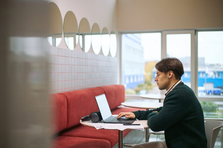 Caucasian young adult man working on laptop at table in modern business setting, sitting alone with...