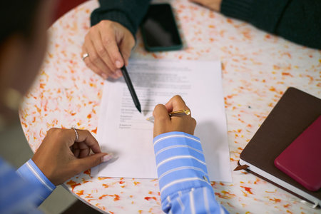 Caucasian woman and Black woman discussing business contract at round table, both reviewing printed document with pen and smartphone visible, focusing on agreement detailsの写真素材