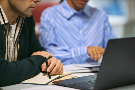Caucasian young adult man and Black young adult woman collaborating at desk, reviewing documents and taking notes while working on laptop in modern business environmentの写真素材
