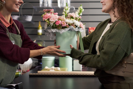 Caucasian young adult woman handing pastel green box, filled with assorted fresh flowers to smiling Caucasian young adult female florist behind counter in flower shop interiorの写真素材