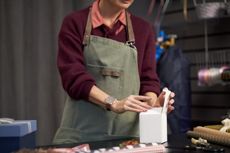 Caucasian young adult woman arranging ribbon on gift box at flower shop counter, hands visible tying...