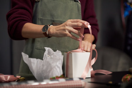 Caucasian young adult woman tying pink ribbon around box while preparing floral arrangement, hands visible with watch on wrist, working at table in flower shop settingの写真素材