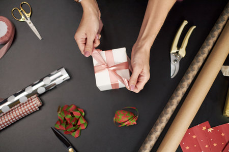 Caucasian middle aged woman tying pink ribbon on gift box surrounded by wrapping paper, scissors, ribbon rolls and holiday decorations on dark table preparing presentの写真素材