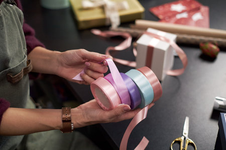 Caucasian young adult woman holding pastel colored ribbons while preparing gift wrapping on black table, hands arranging satin ribbon rolls near wrapped presents and scissorsの写真素材