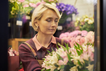 Caucasian middle aged woman arranging fresh flowers in floral shop, focusing on bouquet composition surrounded by various colorful blossoms and green foliage in backgroundの写真素材