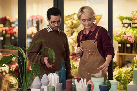 Caucasian middle aged woman arranging flower pots on table beside young adult Black man, discussing floral arrangements in flower shop with colorful bouquets in backgroundの写真素材