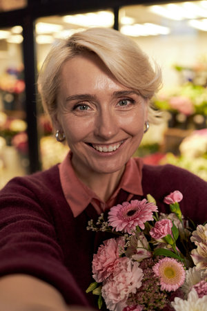 Portrait of middle aged Caucasian woman smiling at camera while holding bouquet of assorted flowers in floral shop, background showing blurred display of various colorful bloomsの写真素材