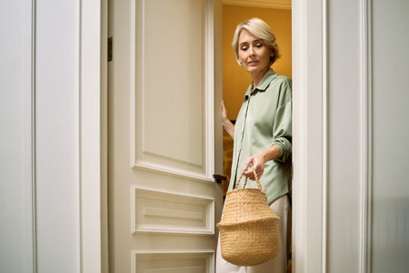Caucasian middle aged woman holding woven basket stepping through doorway, carrying laundry, looking down with relaxed expression, short blond hair visible, indoor residential settingの写真素材