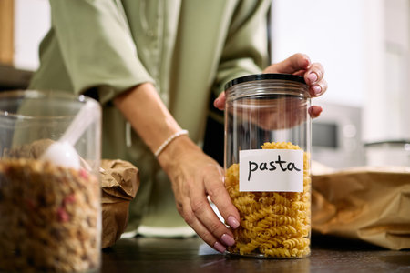 Caucasian young adult woman organizing kitchen pantry, placing transparent container labeled pasta on countertop, hands visible arranging food storage, casual home settingの写真素材