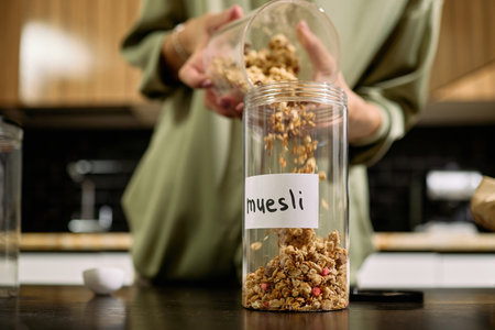 Caucasian young adult woman pouring muesli from plastic container into labeled storage jar in kitchen, hands and torso visible, focusing on food preparation and organizationの写真素材