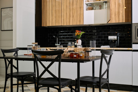 Modern kitchen interior featuring black dining table with empty chairs, countertop displaying various glass jars with food, coffee machine and vase with flowers on backgroundの写真素材