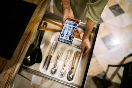 Caucasian young adult woman organizing kitchen utensils in drawer while holding smartphone above tray, capturing photo of neatly arranged cutlery and cooking tools from overhead perspectiveの写真素材