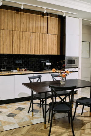 Modern kitchen interior featuring wooden cabinets, black tile backsplash, built-in microwave, dining table with black chairs, glass vase with flowers placed on tabletopの写真素材