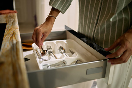Middle aged Caucasian woman organizing silverware in kitchen drawer, hand reaching for spoon in utensil tray, wearing striped shirt, partial body visible, home environmentの写真素材