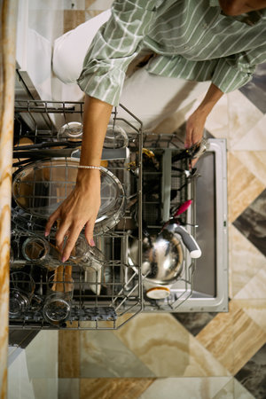 Caucasian middle aged woman loading dishwasher with glassware and kitchen utensils, hand reaching into lower rack, standing on patterned tile floor, partial body visibleの写真素材