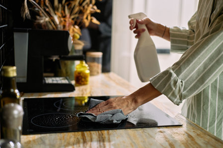 Caucasian middle aged woman cleaning kitchen stovetop using spray bottle and cloth, focusing on wiping surface with hand visible, demonstrating household cleaning routineの写真素材