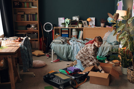 Caucasian teenage girl kneeling on floor packing cardboard box with personal belongings in messy bedroom, suitcase open nearby, engaging in room organizationの写真素材