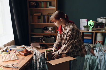teenage girl packing books into cardboard box in bedroom, standing by cluttered desk with various school supplies and personal items, bookshelf in backgroundの写真素材
