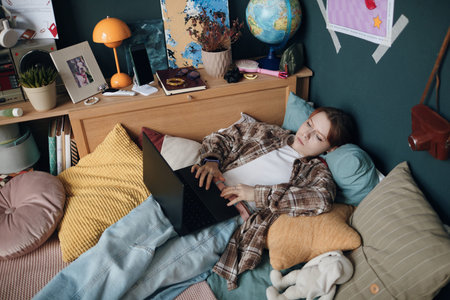 Teenage girl lying on bed using laptop in bedroom surrounded by pillows and personal items, engaging...