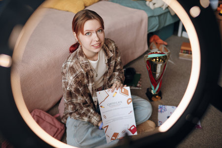 Portrait of Caucasian teenage girl sitting on floor in bedroom holding certificate and looking into camera, trophy and study materials scattered nearby, demonstrating academic achievementの写真素材