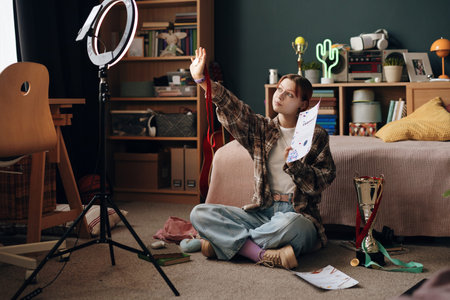Caucasian teenage girl sitting on floor in bedroom recording video with smartphone on tripod ring...