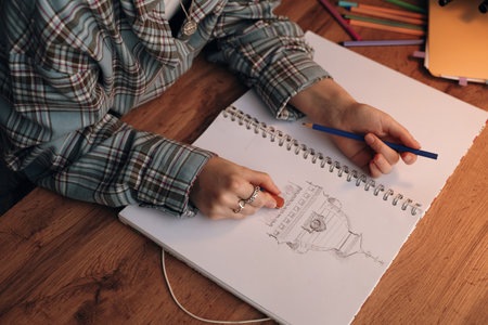 Caucasian teenager drawing architectural sketch in notebook using colored pencil, sitting at wooden desk with colored pencils and digital tablet nearby, focusing on creative activityの写真素材