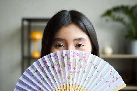 Portrait of young Asian woman looking into camera holding decorative folding fan partially covering face in modern office setting with blurred background elements visibleの写真素材