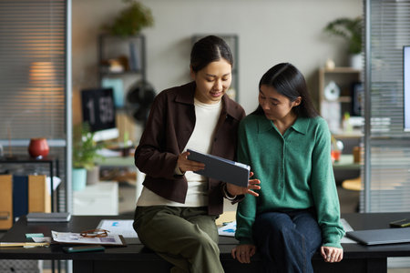 Two young adult Asian women collaborating in modern office, standing and sitting on desk while discussing information displayed on digital tablet, engaging in teamwork and communicationの写真素材