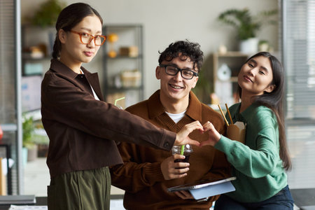 Young Asian man and two young Asian women standing together in office, smiling and forming heart shape with hands, holding drinks and documents, collaborating during work breakの写真素材