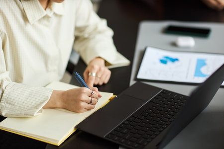 Young Asian woman writing in notebook while working at desk with laptop and digital tablet, focusing on business tasks in modern office environment, hands visibleの写真素材