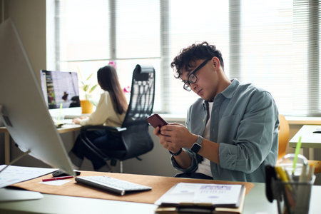 Young Asian man sitting at desk using smartphone while young Asian woman working at computer in modern office workspace, both focused on business tasks and technologyの写真素材