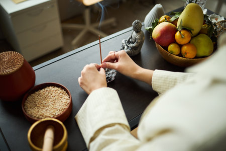 Young Asian woman arranging incense sticks on office desk, hands visible near bowl of fruit and decorative objects, performing mindful ritual during workday in modern workspaceの写真素材