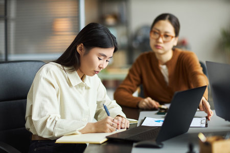 Young Asian businesswomen working together in modern office, one young adult Asian woman writing in notebook while another young adult Asian woman discussing project using laptopの写真素材