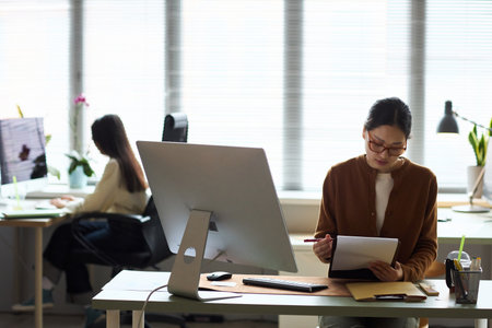 Young Asian businesswoman sitting at desk reviewing documents with pen in hand, large computer monitor in front, another young Asian businesswoman working at computer in backgroundの写真素材