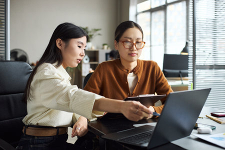 Young adult Asian women collaborating at office desk, one woman pointing at laptop screen while other woman holding digital tablet, both focused on business project discussionの写真素材