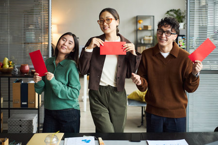Three young adult Asian men and women standing in modern office holding red envelopes, smiling and looking at camera, business documents and laptop visible on desk in foregroundの写真素材
