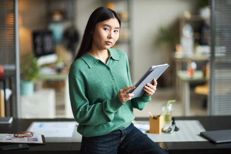 Portrait of young adult Asian woman standing in modern office holding digital tablet, looking confidently at camera, working on business project with documents and office supplies visibleの写真素材